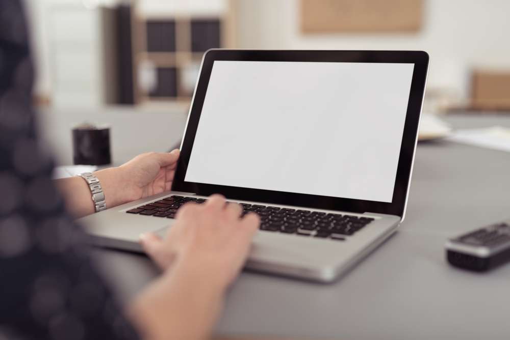 Businesswoman sitting at her desk navigating the internet on a laptop computer using the trackpad, over the shoulder view of the blank screen-1